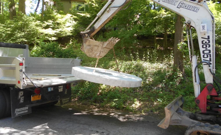 An excavator lifts a round, flat concrete slab with chains above an open dump truck bed on a paved surface.